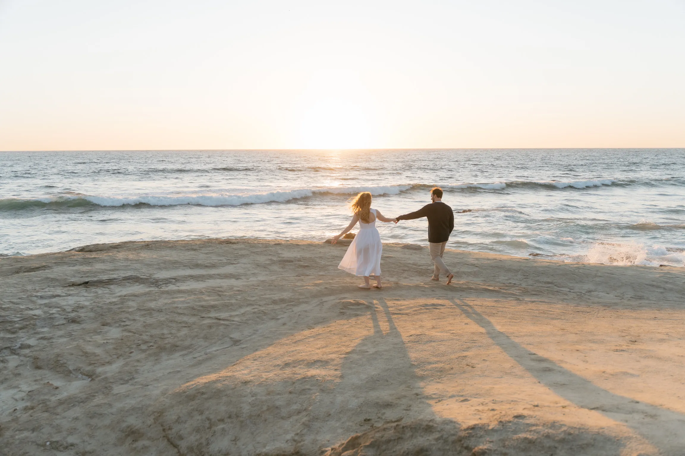 Natalie and Tyler walking hand in hand toward the ocean at sunset.
